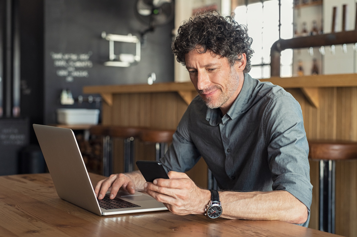 Man with dark curly hair wearing a grey shirt looking at his cell phone while working on his laptop.