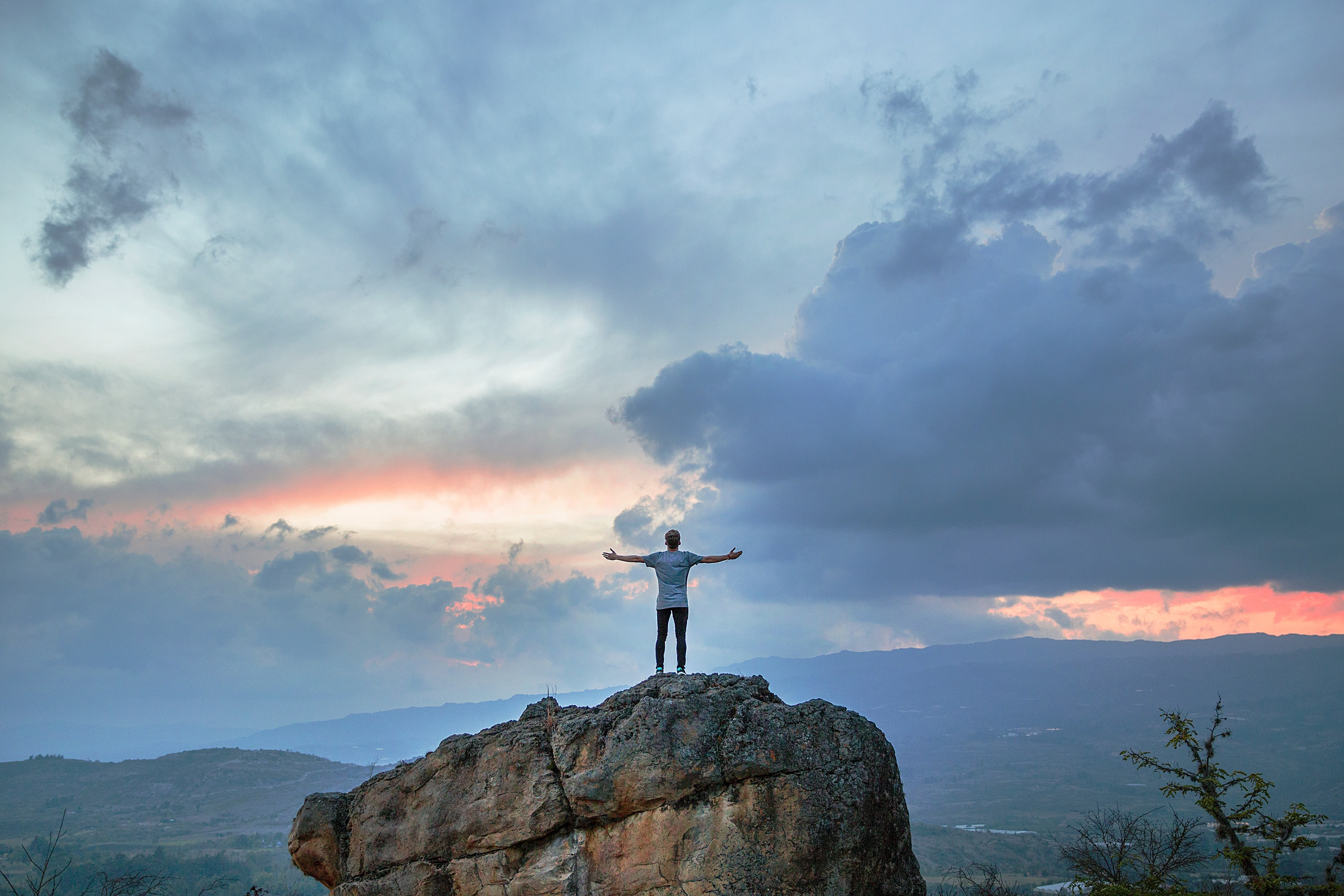 man standing on big rock in a remote area arms outstretched at sunset