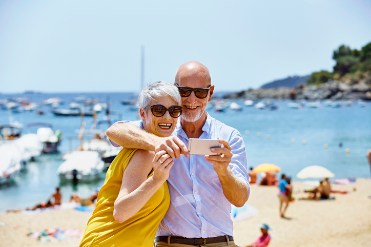 Happy mature couple taking a selfie on the beach with boats behind them.
