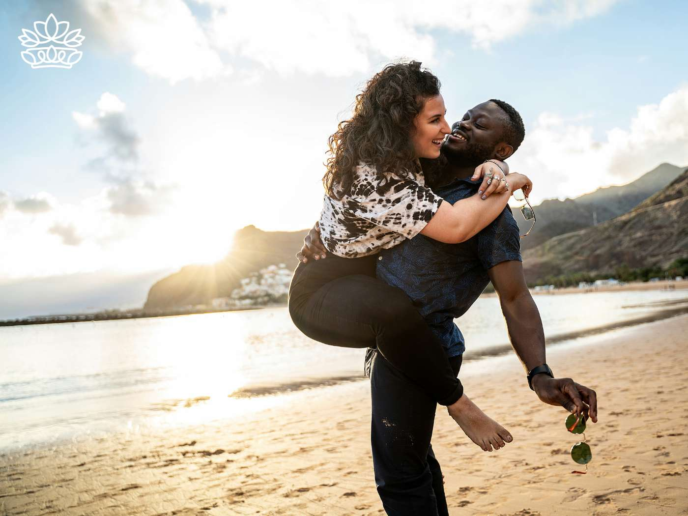 A joyful moment on the beach with a man carrying his partner in a playful embrace, both laughing under the setting sun. This scene epitomizes the happiness and love celebrated by the Engagement Gift Boxes Collection from Fabulous Flowers and Gifts, perfect for such precious life moments.