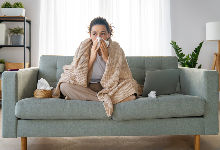 An image of a woman with a cold and throat phlegm sitting on a couch and wiping her nose with a tissue.
