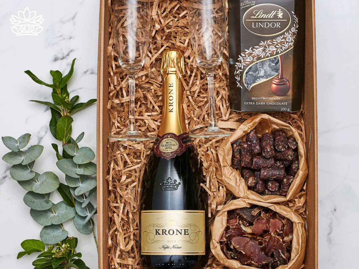 Elegant gift set featuring a bottle of Krone Night Nectar sparkling wine, a crystal flute, Lindt extra dark chocolate, and gourmet dried meats, all beautifully arranged in a wooden crate with natural fill. Graduation. Delivered with Heart. Fabulous Flowers and Gifts.