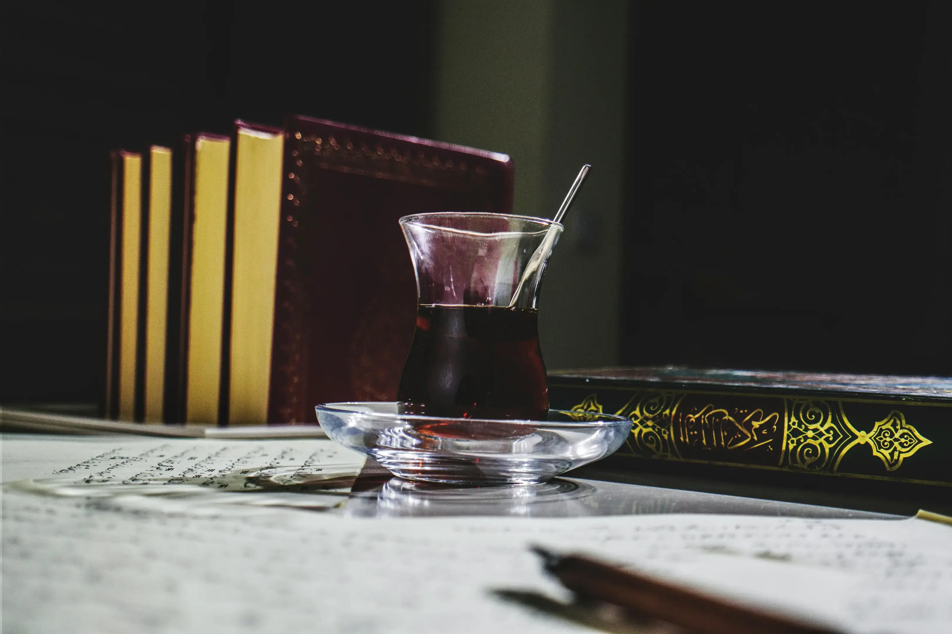 A traditional Turkish tea glass filled with dark tea sits on a clear saucer in the center of a study setup. Surrounding the tea are old-fashioned books, written manuscripts, and a pen under moody, low lighting.