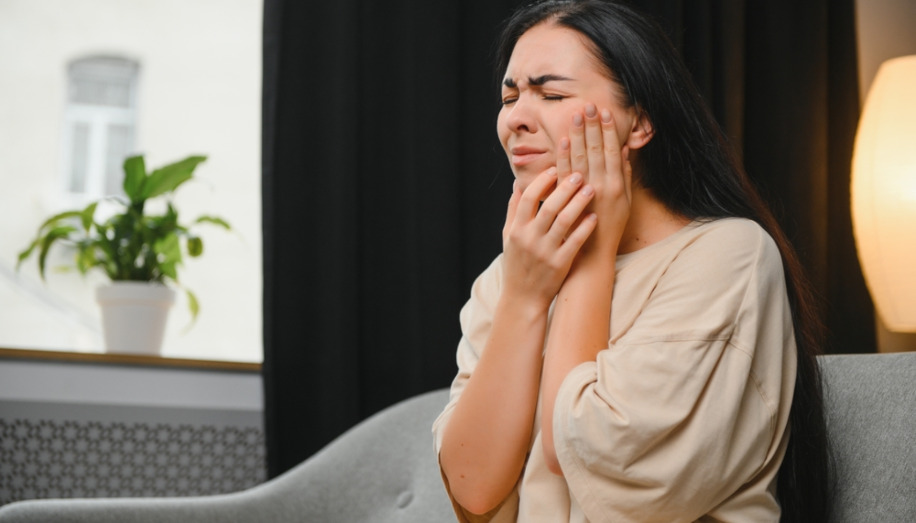 Kvinne sitter på sofa og holder seg til kinnet med smertefullt uttrykk, tydelig plaget av tannpine eller kjevesmerter.