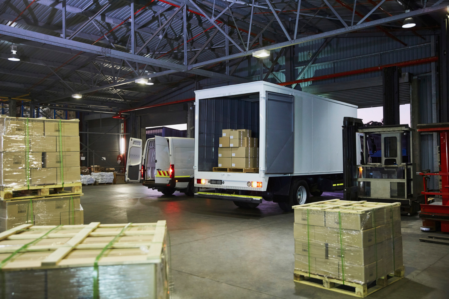 A warehouse with a large truck and a van being loaded with boxes on pallets. A forklift is also visible. It shows how supplement fulfillment possibly happens in a warehouse.