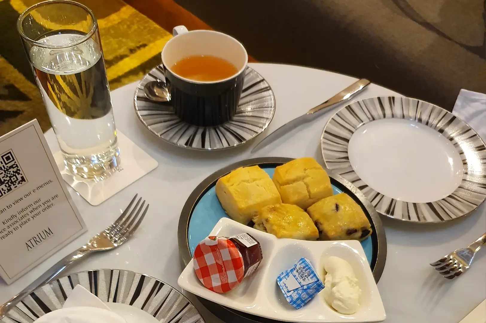 A close-up view of a high tea setting on a white table features a striped teacup and saucer paired with a plate of four golden-brown scones, served alongside a small dish containing a miniature jar of strawberry jam, a butter portion, and a dollop of clotted cream. A tall glass of water and a menu stand with a QR code are also visible on the table, which is set with a silver knife and fork, highlighting the refined details of the afternoon tea service.