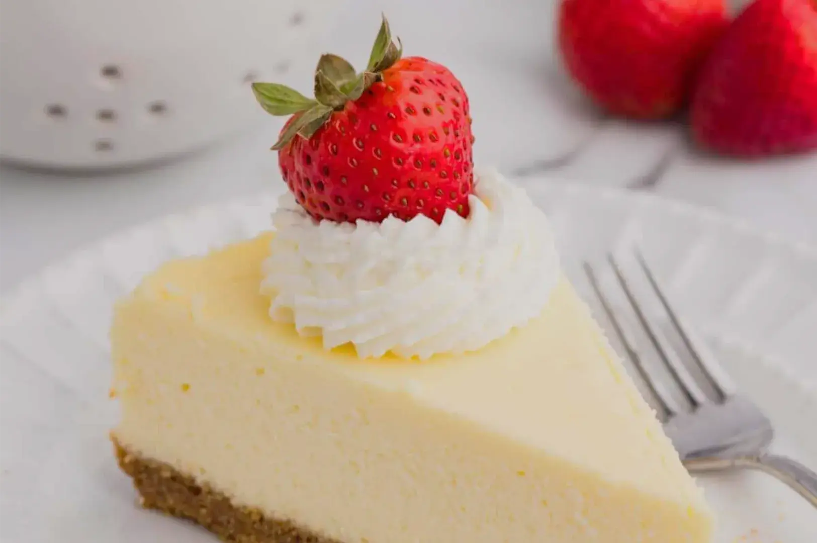 A creamy cheesecake slice with a golden crumb crust is topped with whipped cream and a fresh strawberry, with berries and a fork nearby. The scene feels bright and inviting.