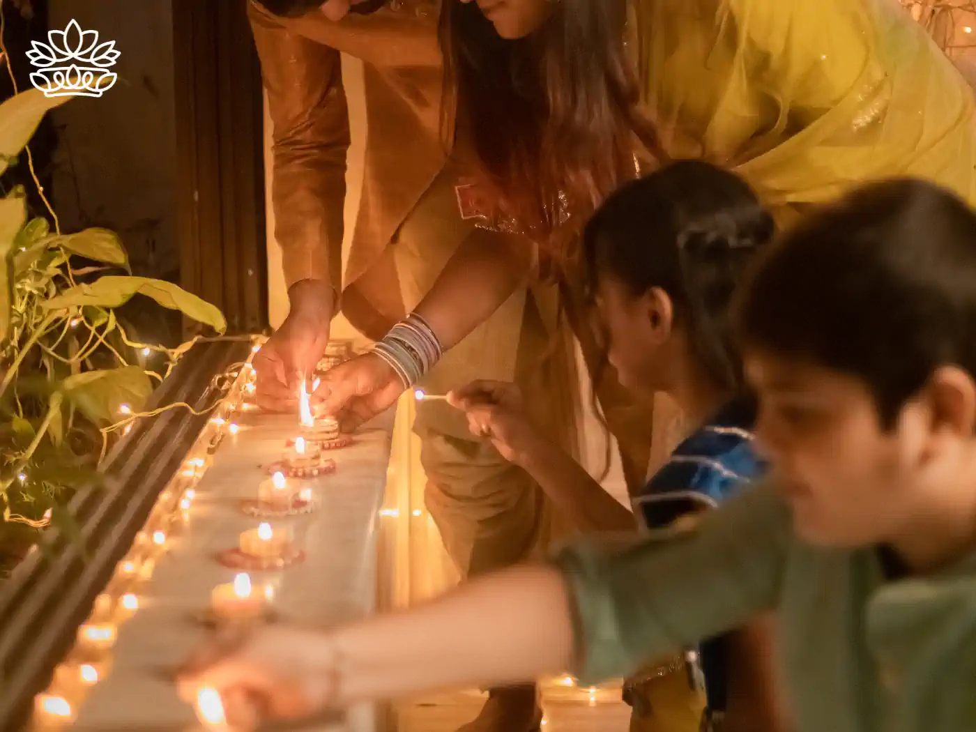 A family engaging in Diwali celebrations, lighting small oil lamps together on a balcony, casting a warm, ambient glow that highlights their festive attire. Fabulous Flowers and Gifts - Diwali Flowers. Delivered with Heart