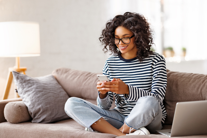A woman with dark hair and glasses sitting on a sofa and smiling at her cell phone.