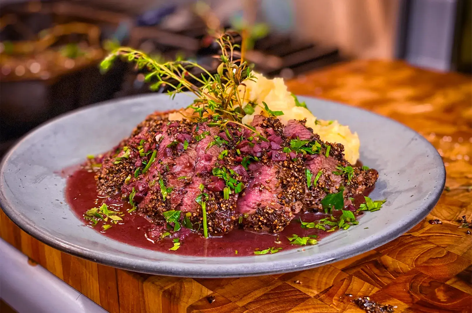 Seared steak crusted with herbs and pepper, garnished with parsley, served with mashed potatoes, on a wooden table with a warm, inviting tone.