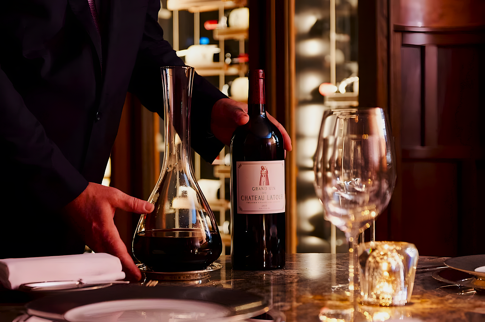 A person in formal attire elegantly holds a large bottle of red wine next to a glass decanter on a restaurant table, surrounded by wine glasses and soft candlelight.