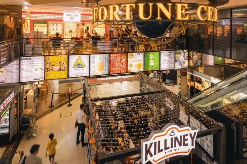 A multi-level interior view of the Fortune Centre atrium, showcasing a Killiney coffee kiosk on the ground floor, brightly lit restaurant signboards on the upper level, and patrons walking through the mall.