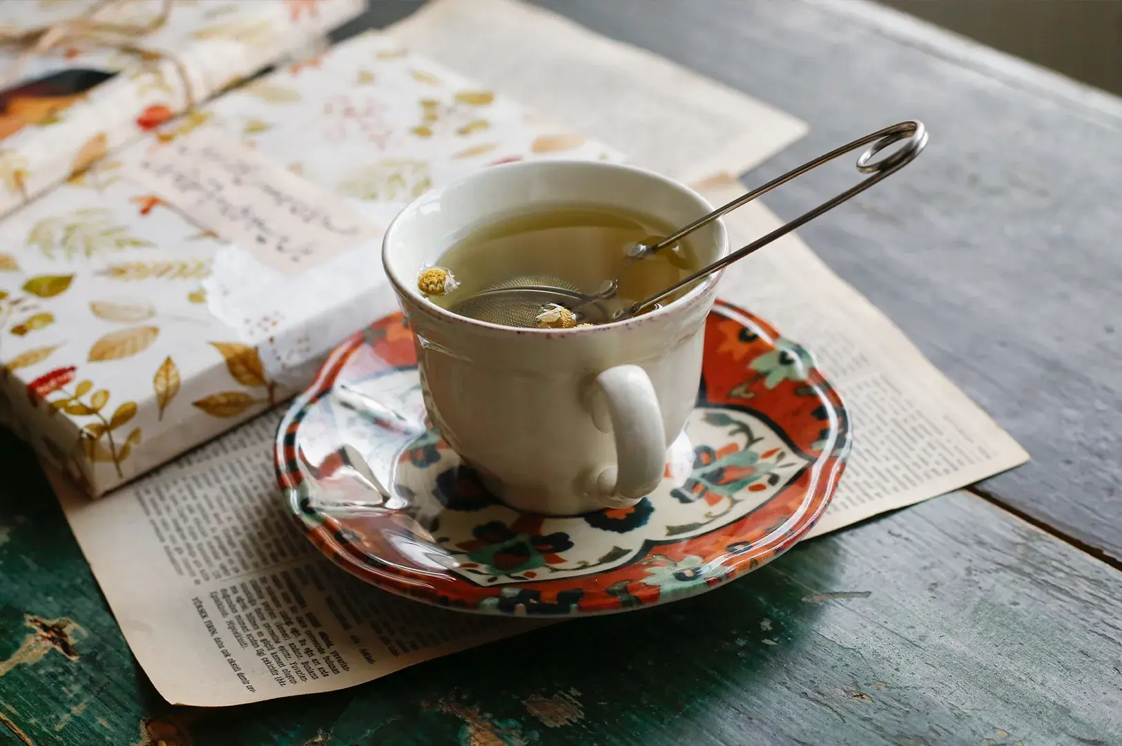 A close-up shot displays a vintage-style white ceramic teacup filled with light herbal tea, resting on an intricately patterned saucer with red, green, and orange floral motifs. Inside the cup, a metal mesh scissor-style tea infuser holds chamomile flowers, with a few small buds floating freely in the liquid. The entire tea set is placed on top of open, aged book pages on a rustic, distressed green wooden table, creating a nostalgic and literary atmosphere.