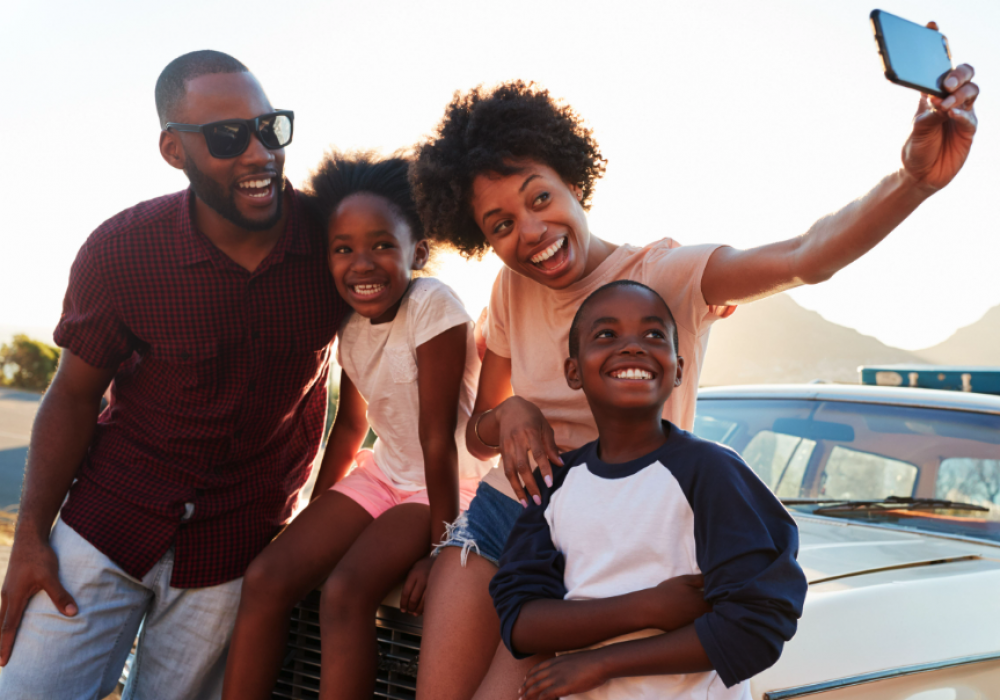 Smiling family sitting on the hood of a car taking a selfie. 
