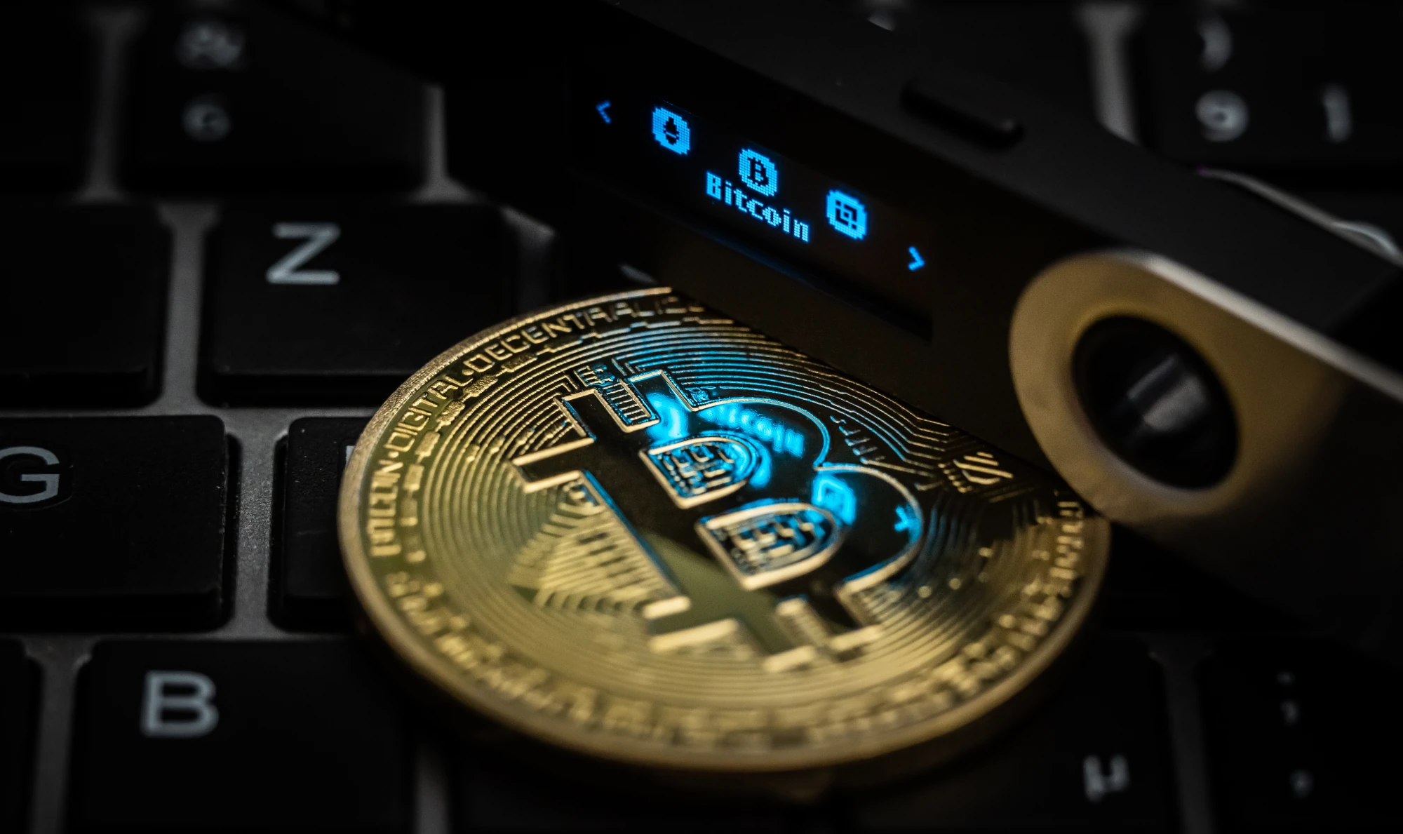 The word Bitcoin can be seen on the display of a Ledger Nano S hardware wallet sitting on top of a symbolic "Bitcoin coin" which is resting on a laptop keyboard.