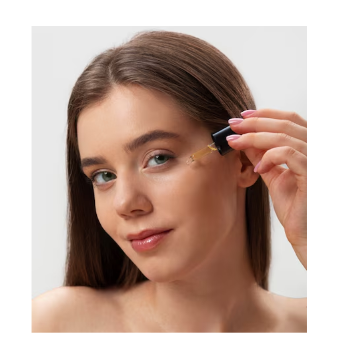 Vrouw met een frisse, egale huid brengt met een pipet een huidverzorgingsserum aan op haar gezicht, net onder het rechteroog. Ze kijkt rustig in de camera, terwijl het transparante serum uit haar skincare routine voorzichtig op haar huid wordt gedruppeld. De setting is helder en minimalistisch, met een focus op skincare en dagelijkse huidverzorgingsroutines. De afbeelding illustreert het gebruik van een gezichtsserum, mogelijk met niacinamide of een vergelijkbaar ingrediënt.