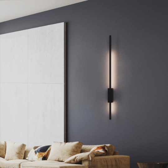 Modern wall-mounted vertical light fixture emitting a soft glow, installed on a dark accent wall above a neutral-toned couch with plush cushions in a contemporary living room.