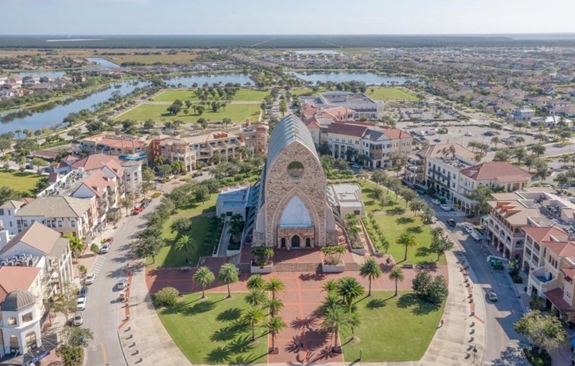 An aerial view of the Ave Maria master-planned community showcases its residential neighborhoods and the vibrant town center, featuring resort-style amenities such as a championship golf course and a fitness center. The image highlights thoughtfully designed living spaces, including homes with spacious kitchens and outdoor areas, all set against a serene backdrop in Southwest Florida.