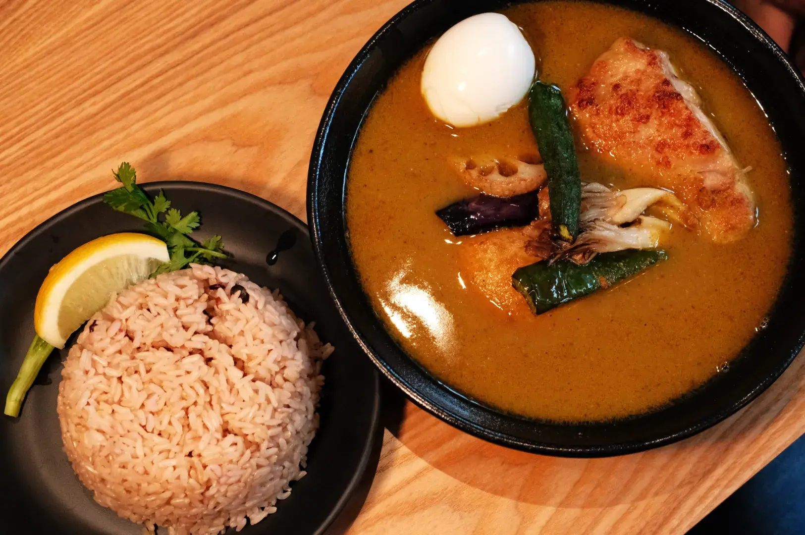 A bowl of curry with chicken, boiled egg, and vegetables, served with a side of brown rice and a wedge of lemon on a black plate.