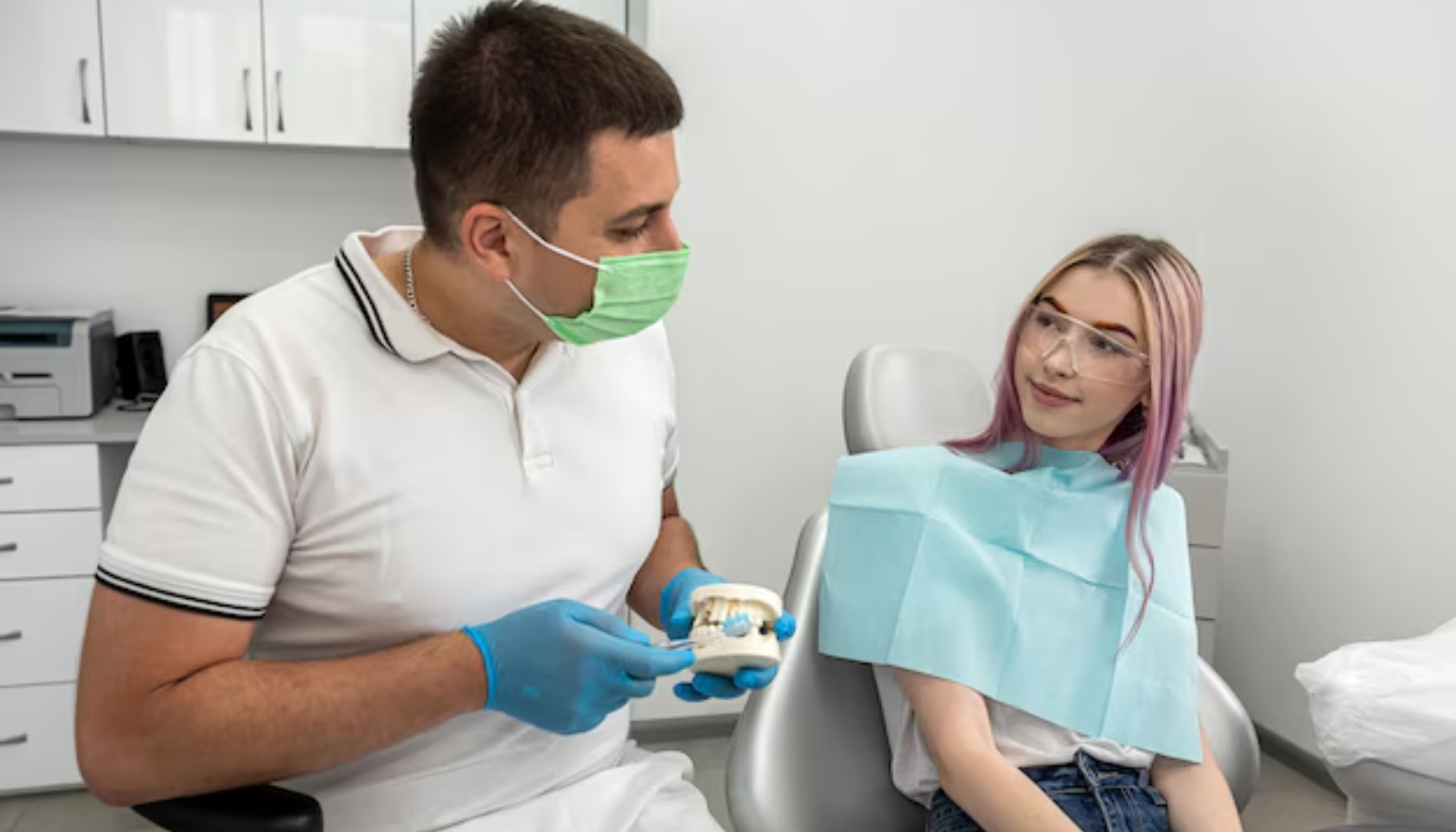 Tannlege med grønt munnbind og hansker forklarer en pasient behandling ved hjelp av en tannmodell.