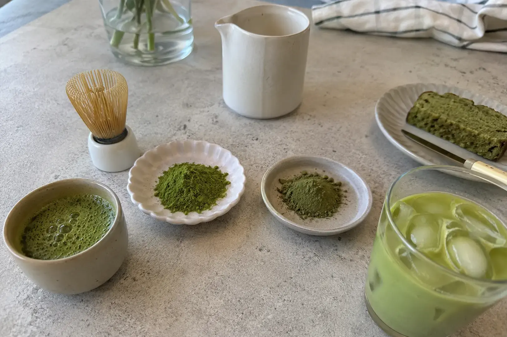 Presents a bright, clean kitchen workspace featuring two small white dishes filled with different shades of powdered green tea, a traditional bamboo matcha whisk, a white ceramic pitcher, and a glass of iced matcha, accompanied by a small plate of sliced green tea bread.