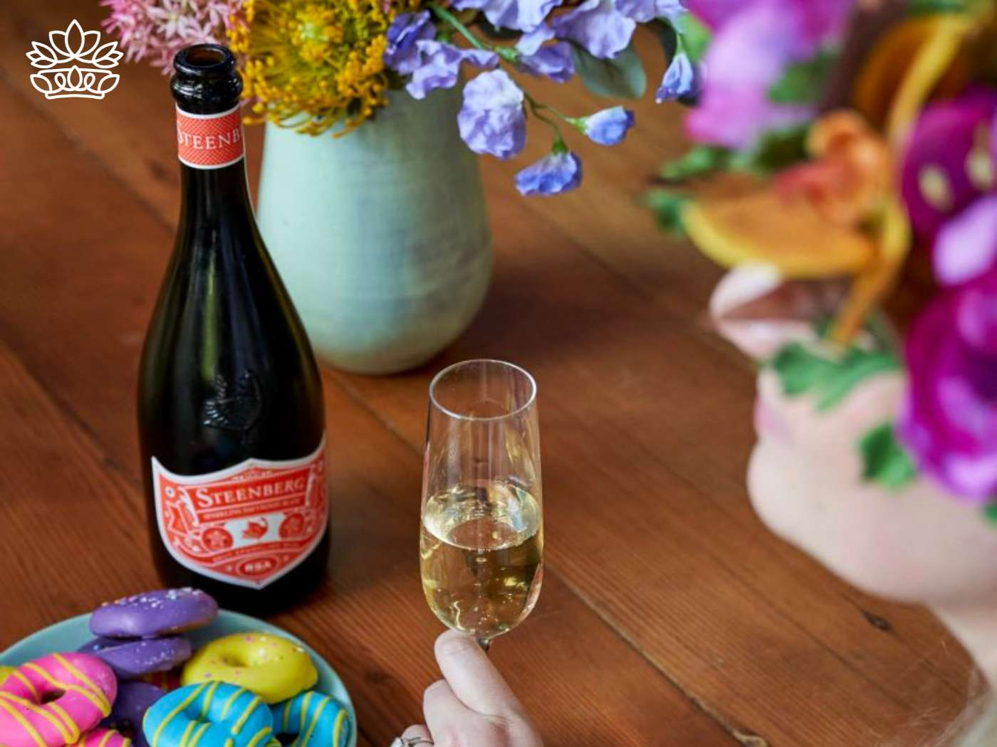 A hand holding a glass of sparkling wine beside a bottle, with vibrant macarons on a plate and a bouquet of fresh flowers in a vase, celebrating with the Congratulations Messages —Fabulous Flowers and Gifts.