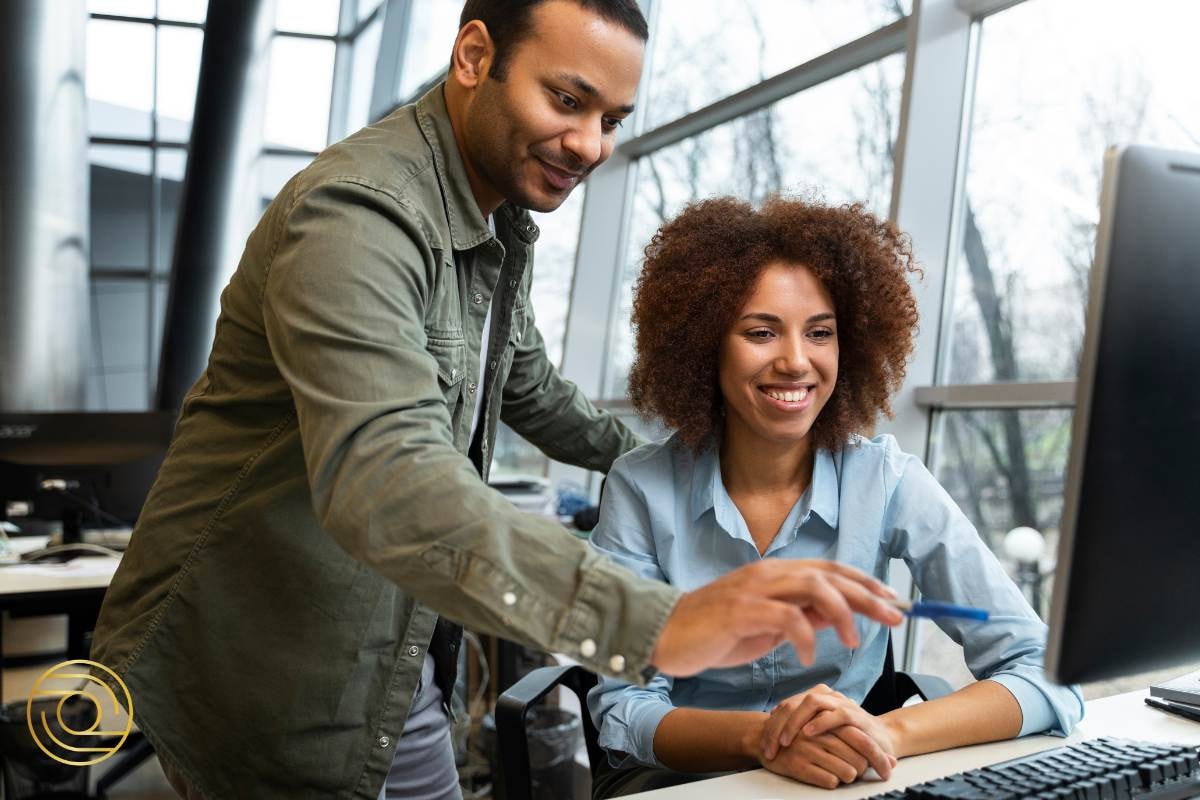 Two colleagues working together at a computer in an open office space, with one pointing at the screen and the other smiling.