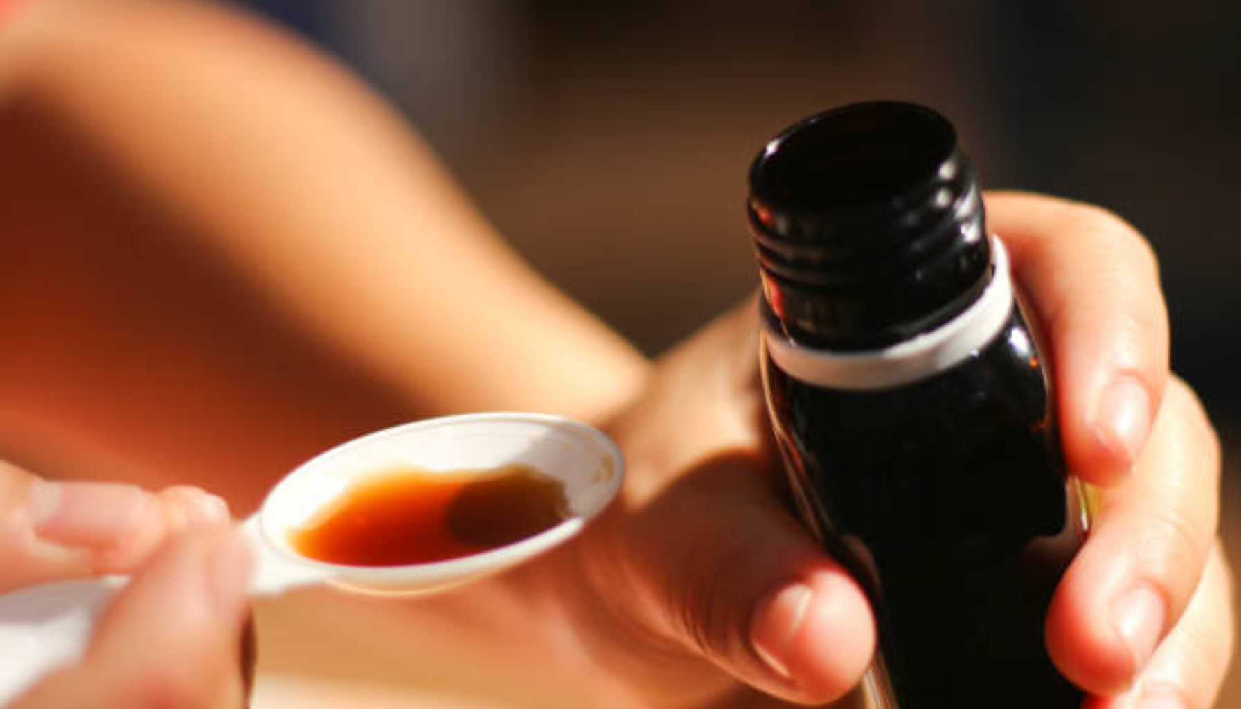 Person heller flytende medisin i en plastskje fra en mørk flaske, klar til inntak.