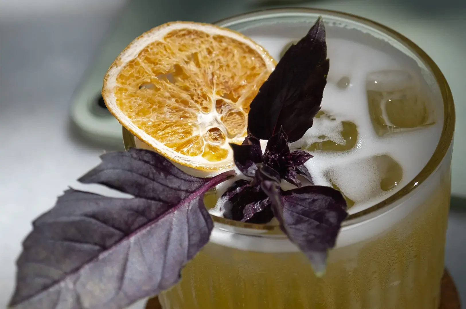 Close-up top-down view of an iced cocktail garnished with a dried citrus wheel and deep purple basil leaves.