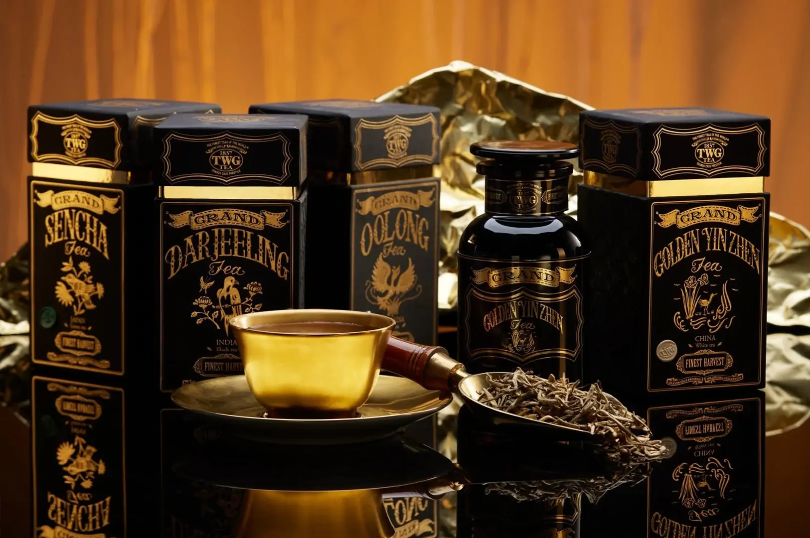 A collection of high-end tea boxes from TWG Tea arranged on a reflective black surface. The boxes are black with ornate gold lettering and borders. Prominently featured are "Grand Sencha," "Grand Darjeeling," "Grand Oolong," and "Grand Golden Yin Zhen." A small gold cup and saucer sit in front, alongside a golden scoop filled with loose white tea leaves. The background is a warm, metallic gold tone.