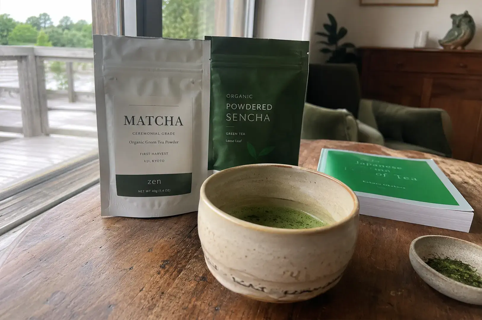 A rustic wooden table serves as a setting for a bowl of prepared green tea, positioned beside two pouches—one for ceremonial grade matcha and one for organic powdered sencha—and a copy of the book The Japanese Tea of Tea by Kakuzo Okakura.