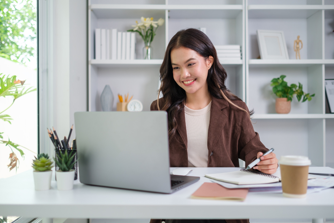 Filipina virtual assistant smiling while working on her laptop with notebooks and coffee on desk