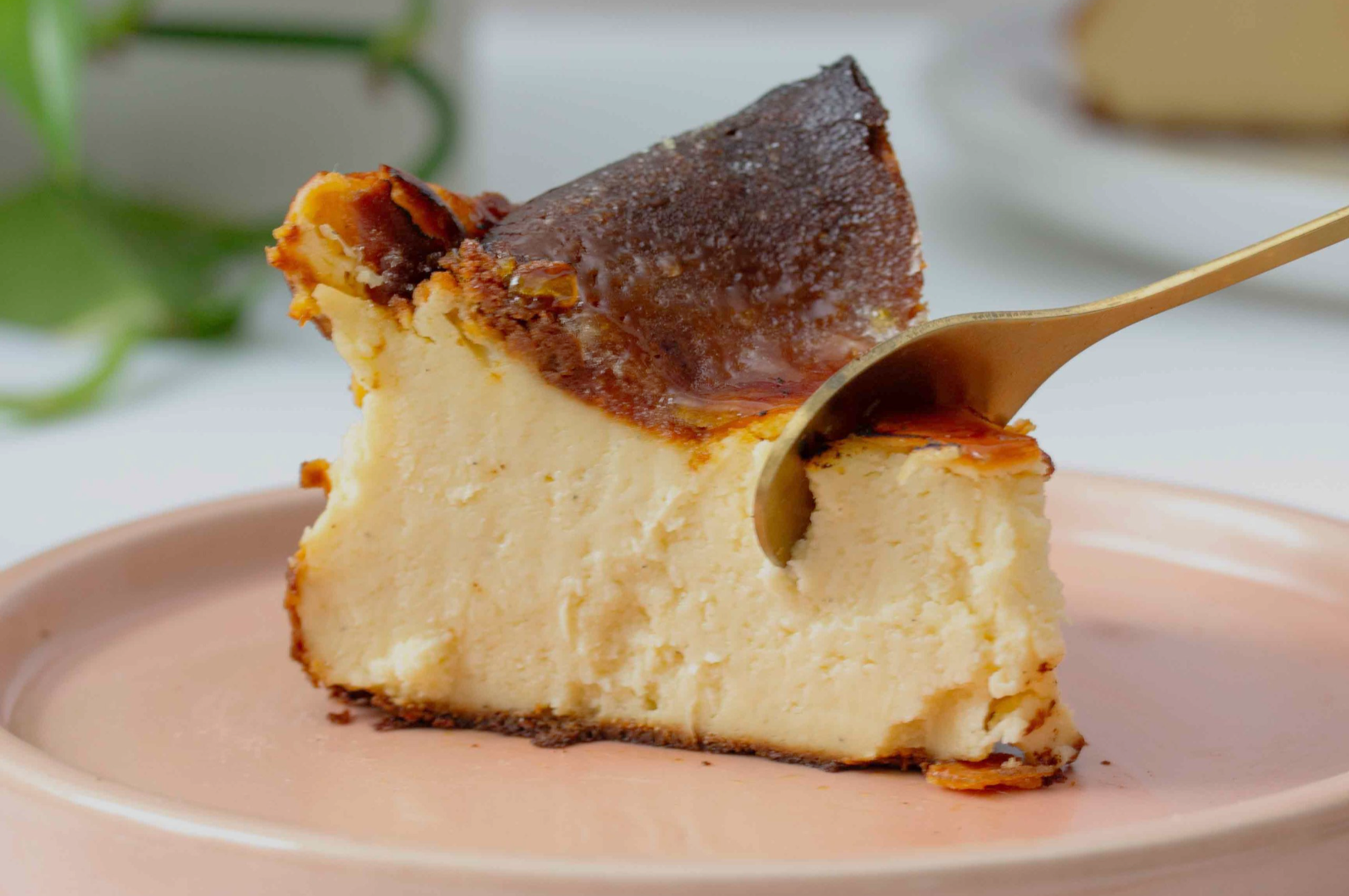 A slice of creamy cheesecake with a caramelized top on a pink plate. A spoon gently presses into its soft texture, evoking a sense of indulgence.