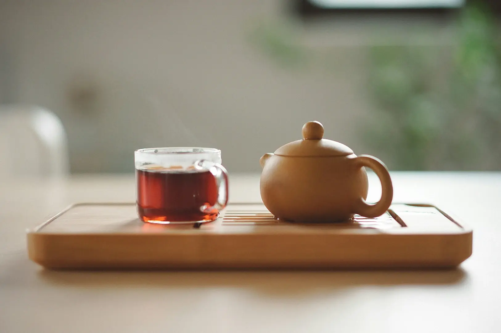 A simple and serene tea setup on a light-colored wooden table. A small, round, unglazed brown clay teapot sits beside a small, clear glass cup filled with amber-colored tea. The items are arranged on a bamboo tea tray, and the background is softly out of focus.