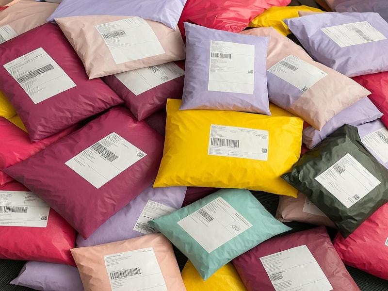 A large pile of colorful plastic shipping packages. The packages come in various colors, including pink, purple, yellow, black, and beige. Each package is sealed and has a white shipping label with barcodes and addresses printed on it. The labels indicate that these packages are ready for delivery.