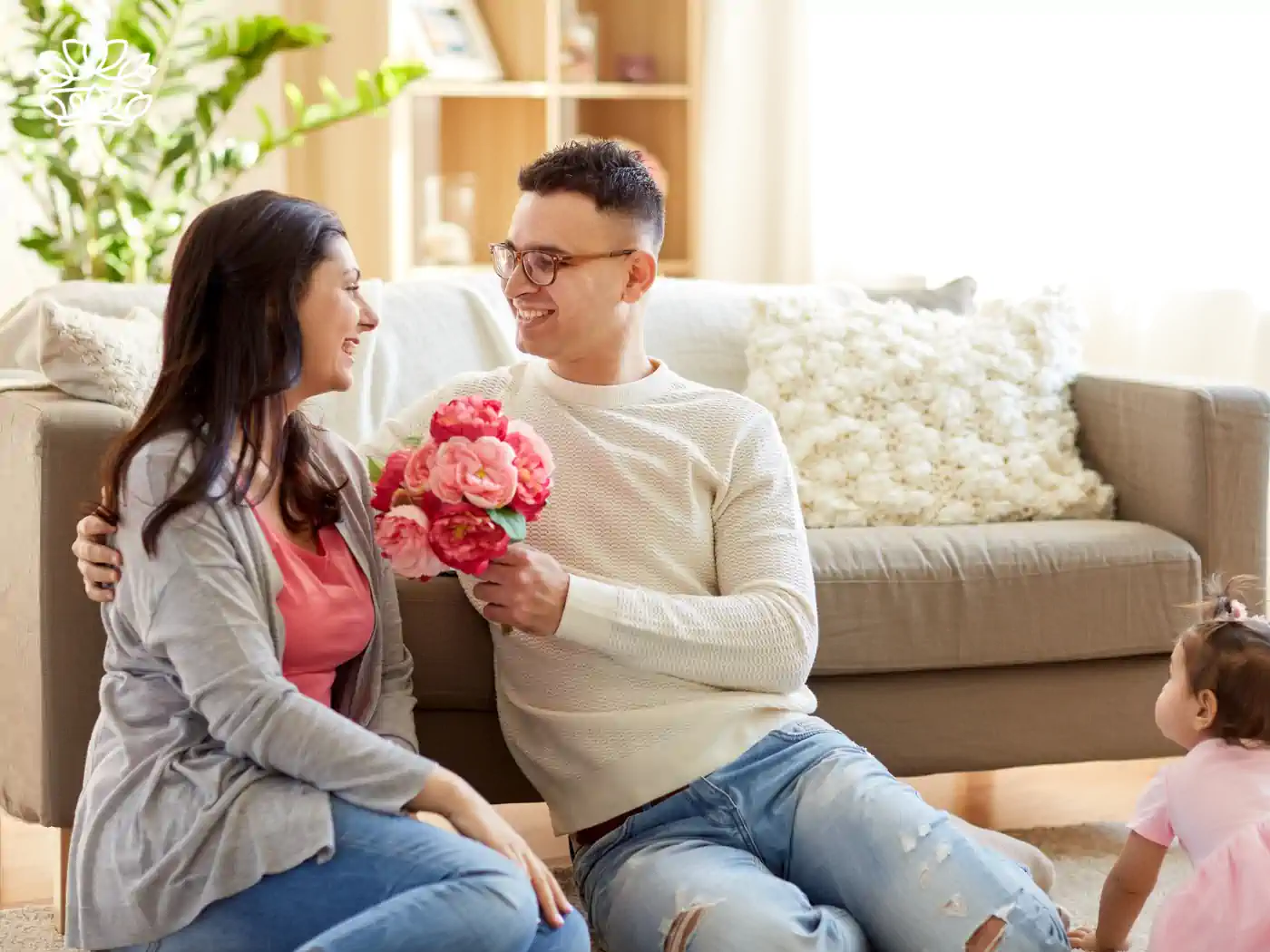 A smiling man sitting on the floor presents a bouquet of pink peonies to his delighted wife, with their young daughter watching in a warmly lit living room. Just Because Flowers. Delivered with Heart. Fabulous Flowers and Gifts.