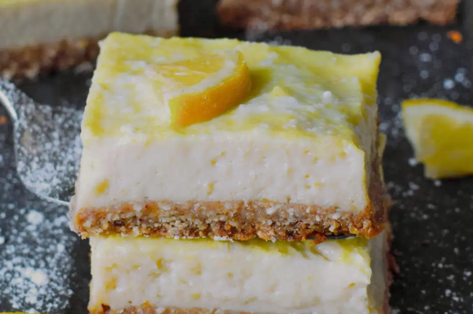 A stack of lemon cheesecake squares with a slice of lemon on top and a crumbly base.
