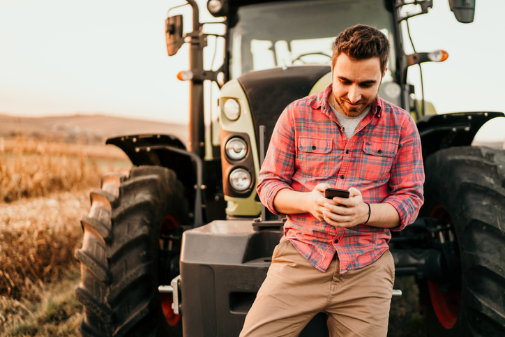 Happy young man in a red plaid shirt leaning against a tractor and checking his email.