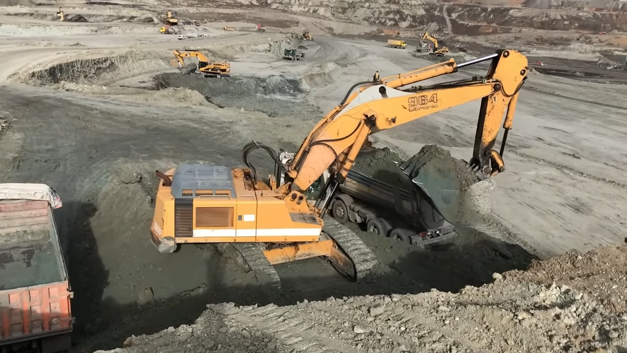 A large orange excavator is scooping up soil and loading it into a dump truck in an open-pit mine, with several other construction vehicles visible in the background