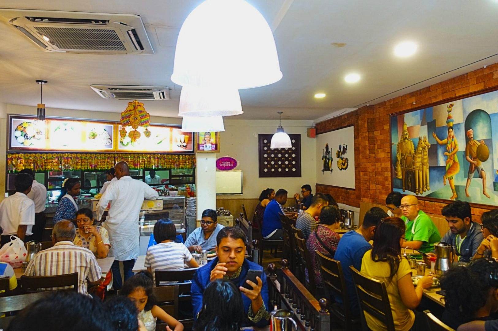 A bustling Indian restaurant with customers seated at tables, enjoying meals. Bright lighting and colorful wall art create a vibrant, lively atmosphere.