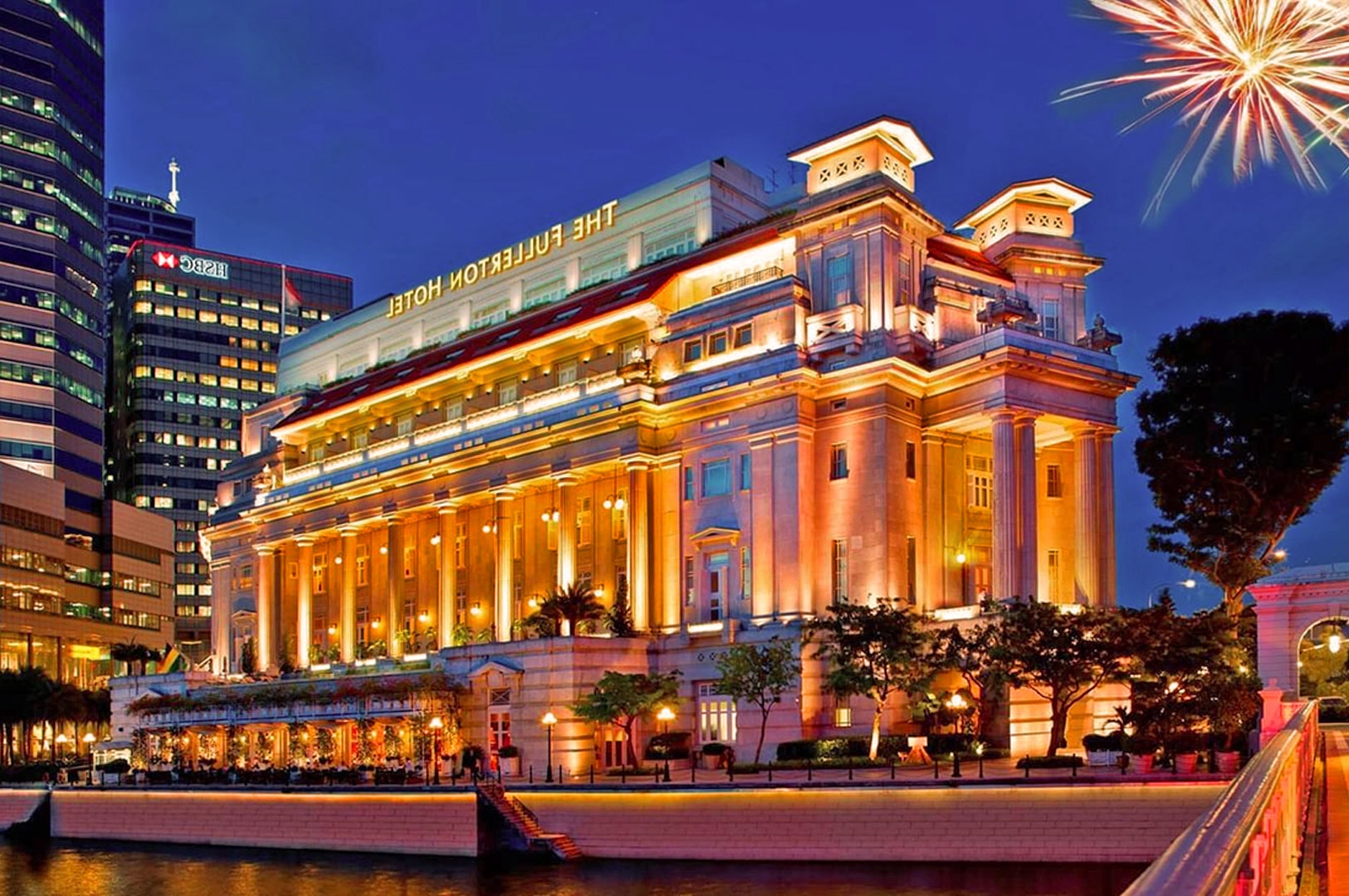 Illuminated grand hotel at night, reflecting on a river. Fireworks burst in the sky, creating a festive ambiance. Tall buildings in the background.