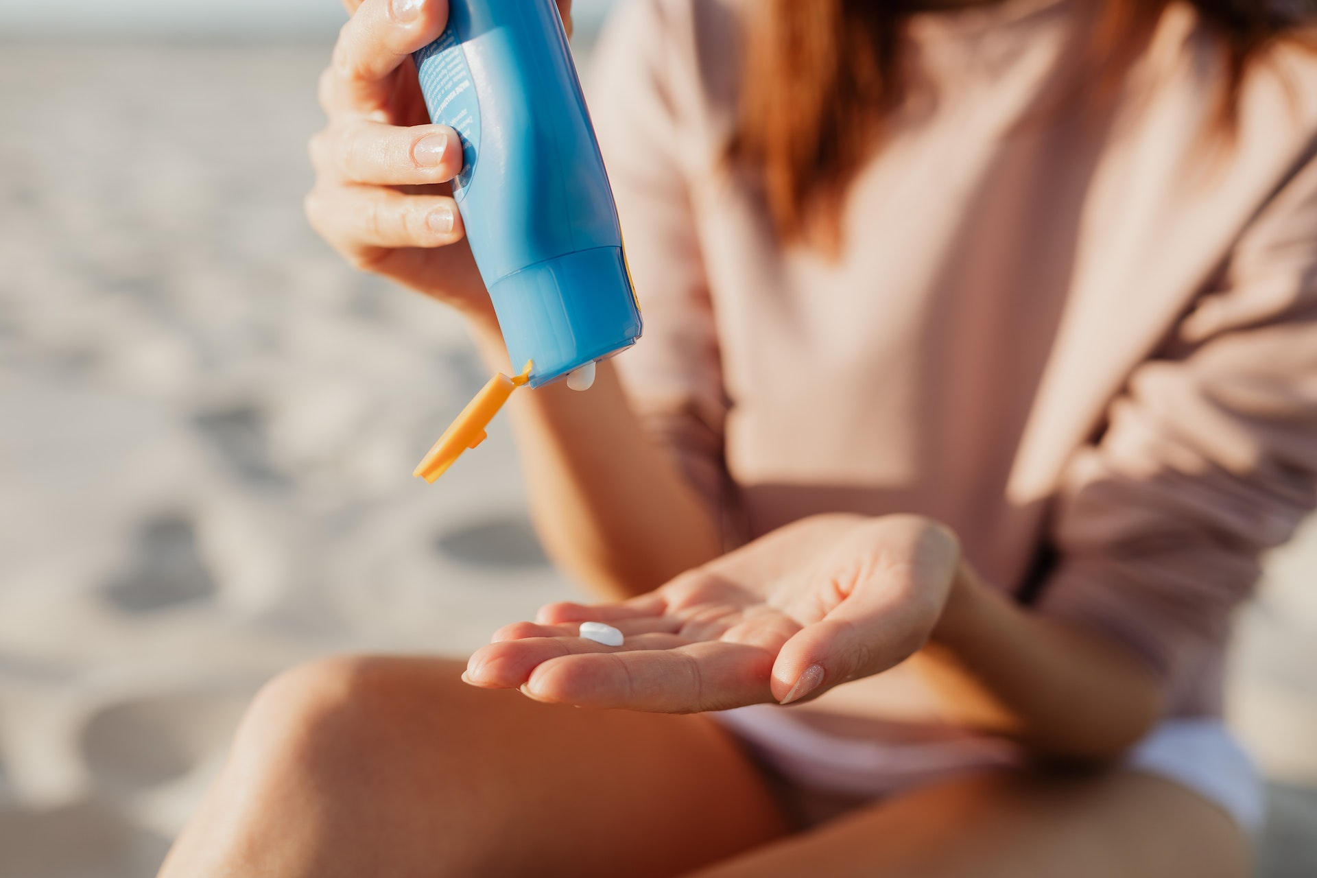 Jovem sentada na praia derramando o protetor solar em uma das mãos. Não é possível ver seu rosto. Ela usa um moletom leve, de mangas compridas, e um shorts branco. Imagem - Pexels.com