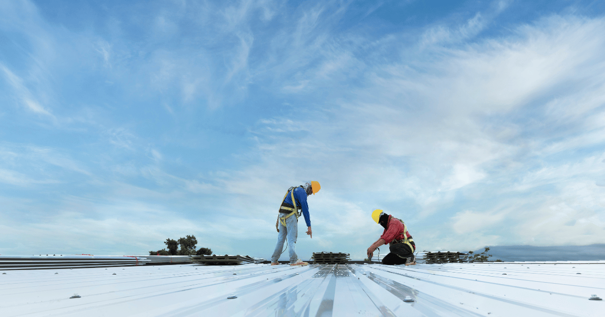 Two roofers in safety gear positioning metal panels on a commercial roof against a backdrop of a blue sky.