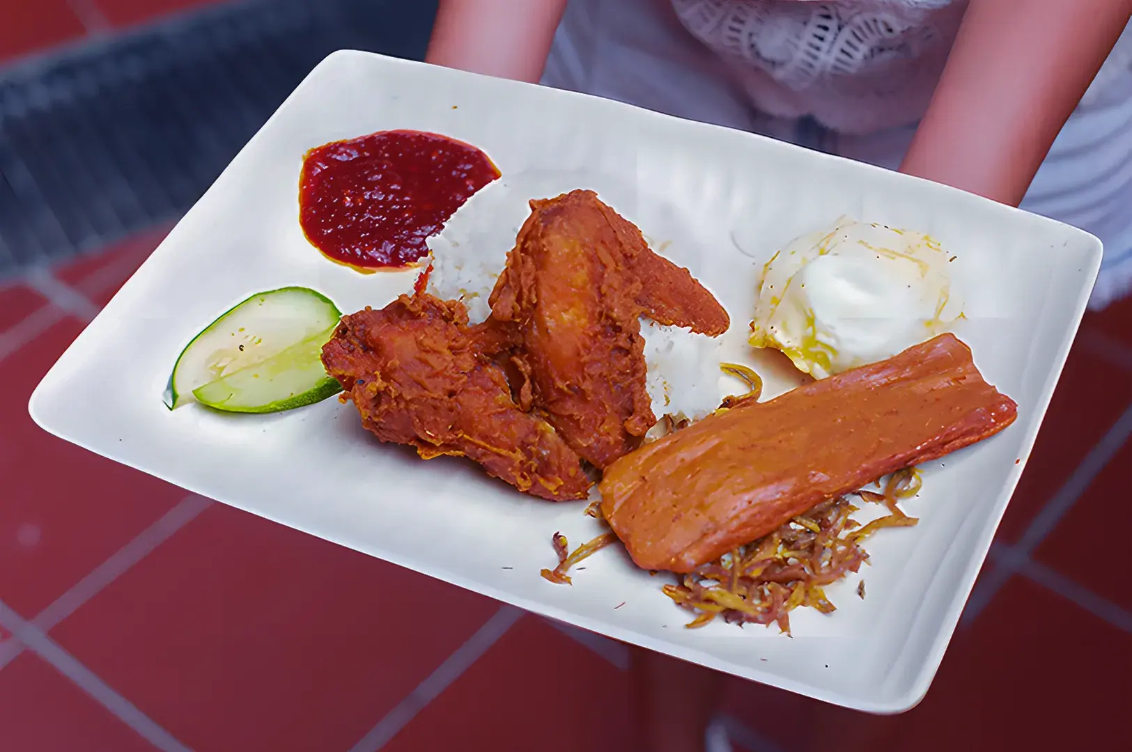 A plate of nasi lemak with fried chicken, sambal, cucumbers, anchovies, a poached egg, and a slice of luncheon meat, held on a red-tiled floor.