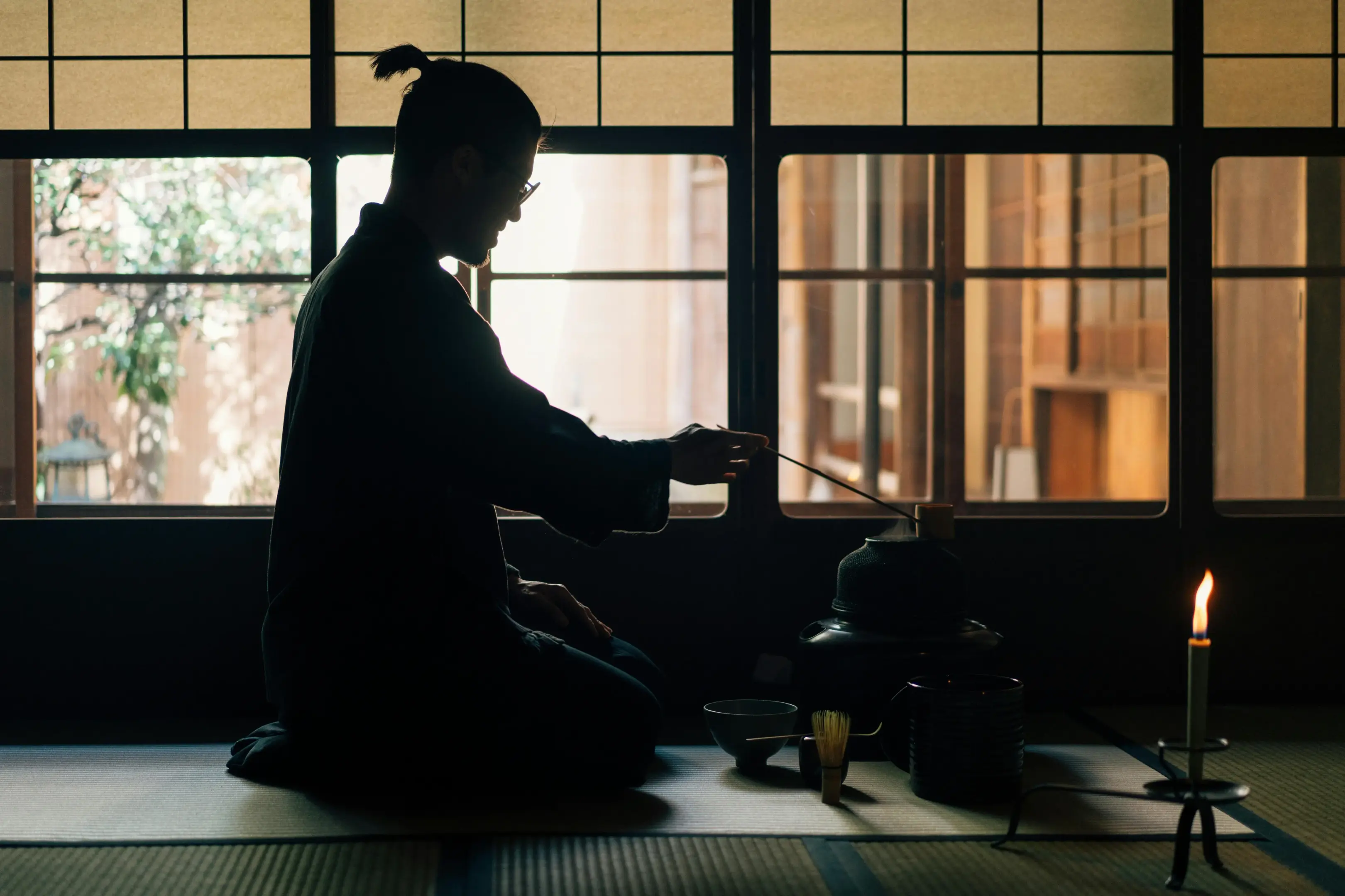 A man sits in silhouette within a traditional Japanese room, meticulously performing a tea ceremony before a lit candle and steaming kettle. Warm light filters through shoji screen windows, casting a serene glow over the tatami mat and tea utensils.