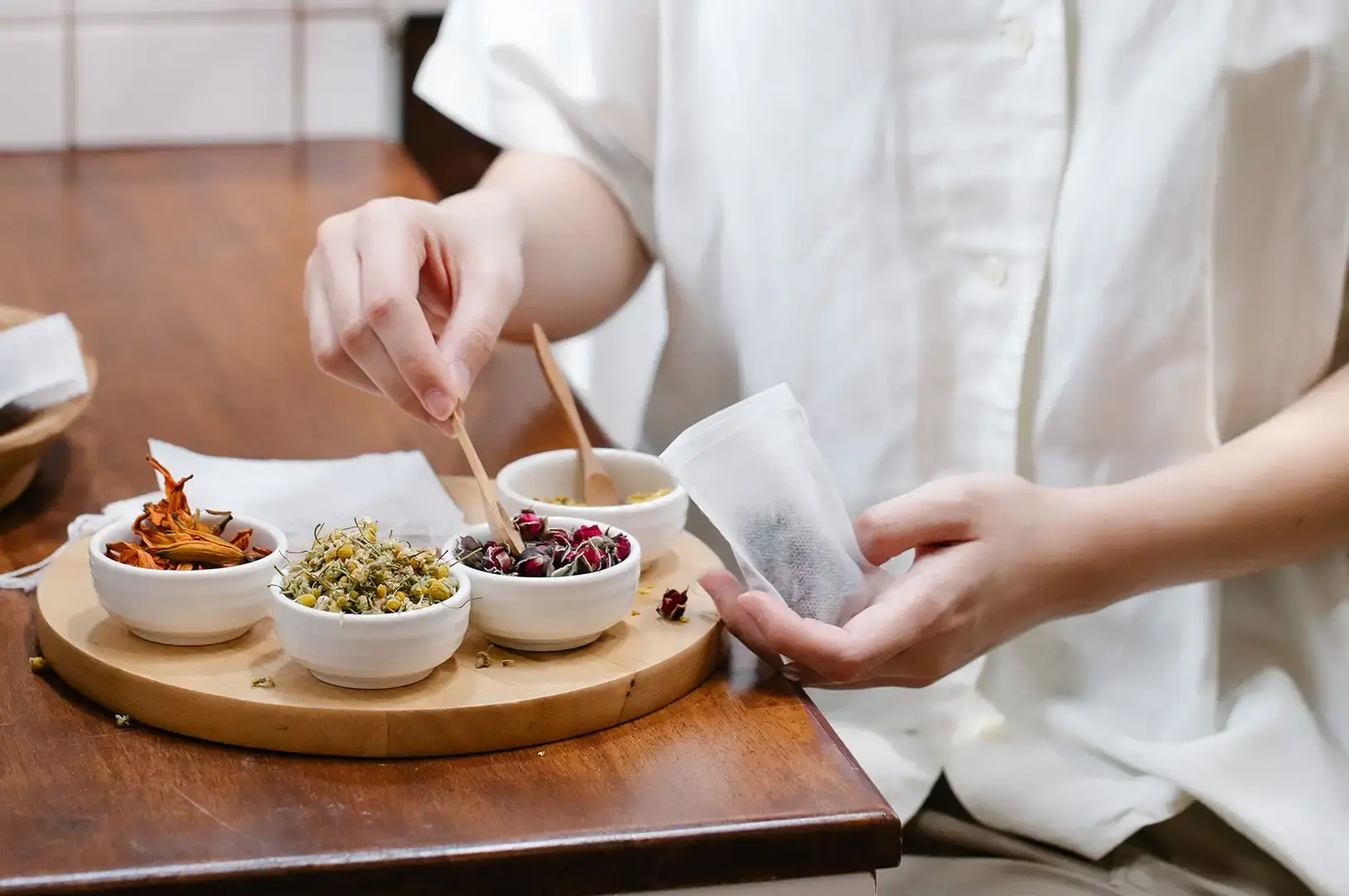 A person dressed in a white linen shirt using a small wooden spoon to scoop dried rosebuds into an open white filter tea bag, with a round wooden tray holding small white bowls of various dried herbs and flowers arranged on a dark wooden table.