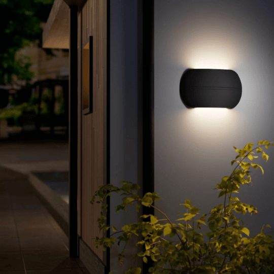 Modern oval-shaped outdoor wall light emitting a soft up-and-down glow, mounted on a white wall near plants in a nighttime setting.