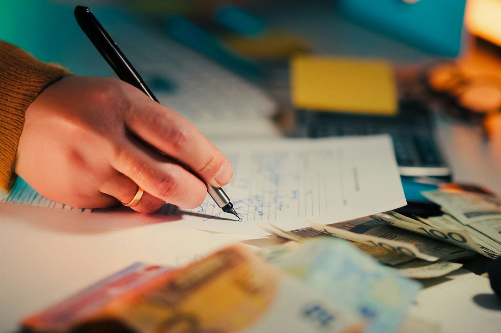 A hand holding a pen writes on a document, surrounded by euro banknotes and a calculator. The scene conveys a focused, financial planning atmosphere.