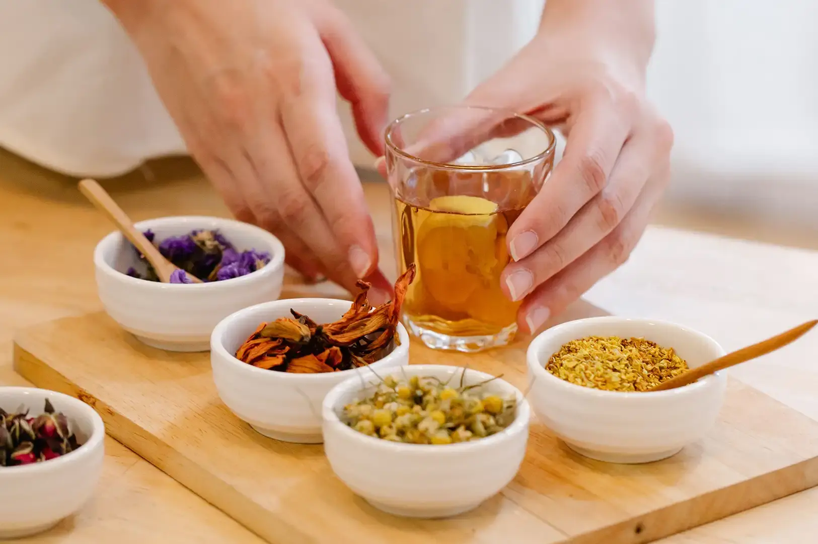 A person's hands framing a clear glass filled with freshly brewed warm amber tea on a light wooden board, surrounded by several small white ceramic bowls holding vibrant dried botanical ingredients including purple blossoms, orange lily petals, chamomile, and yellow osmanthus flowers.