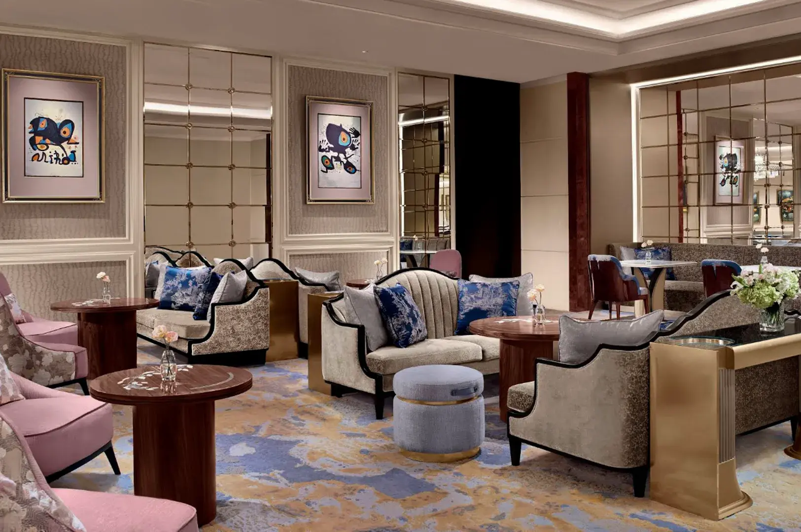 This wide shot captures an elegant and luxurious hotel lounge decorated in sophisticated muted tones, featuring plush seating arrangements spaced across a large, patterned carpet with swirling shades of blue, gold, and cream. The main seating area includes curved beige sofas adorned with deep blue and silver-patterned throw pillows, paired with small round wooden coffee tables, a low grey velvet ottoman with a gold base, and armchairs with soft pink velvet upholstery. The walls are finished in a neutral textured wallpaper, punctuated by large grid-patterned mirrors that reflect the room and modern abstract framed artwork, creating an inviting yet upscale ambiance.